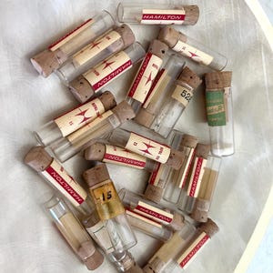 May include: A collection of small, clear glass vials with cork stoppers. Many vials have labels with the word "HAMILTON" in red. Some labels also include numbers and the word "REFILL". The vials are arranged in a circular pattern.