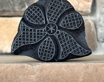 May include: Black wooden block with a carved design of a flower with a grid pattern. The flower has four petals and a center with a star shape.