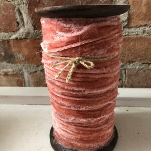 May include: A spool of dusty rose pink velvet ribbon with frayed edges, wrapped with twine and sitting on a wooden base.