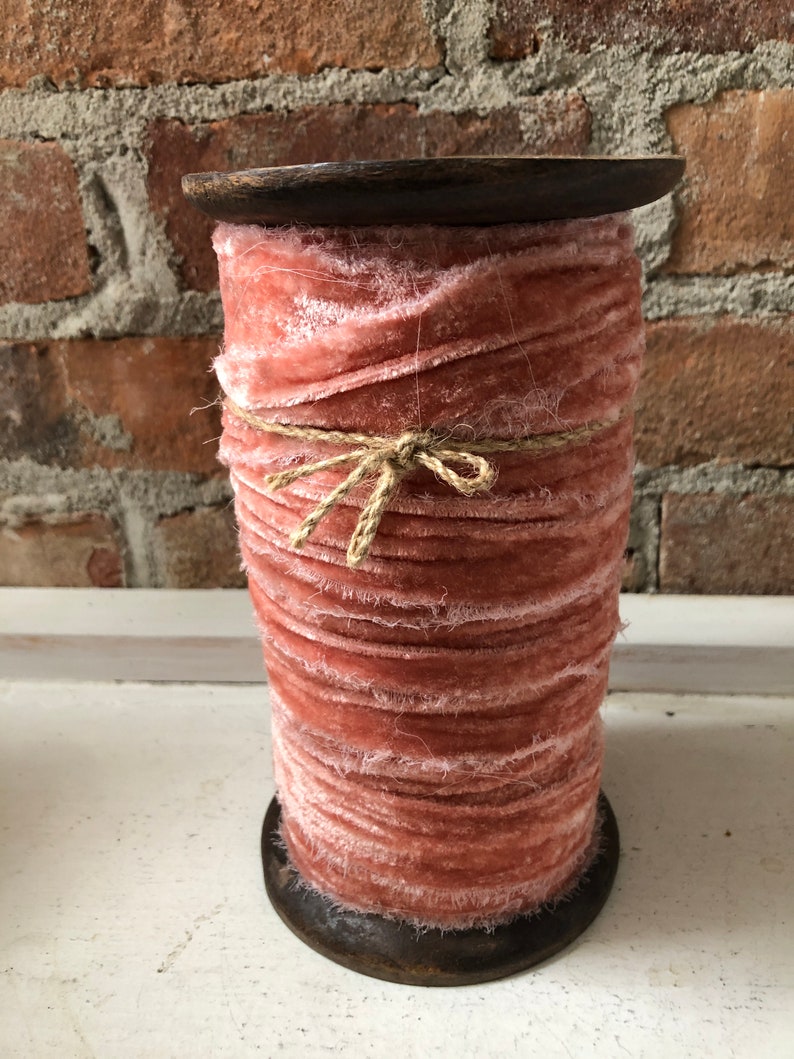 May include: A spool of dusty rose pink velvet ribbon with frayed edges, wrapped with twine and sitting on a wooden base.