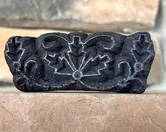 May include: A black wooden block printing stamp with a carved floral design. The design features a central flower with four petals and leaves surrounding it.