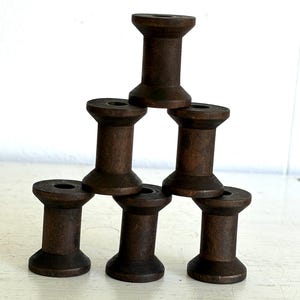 May include: A stack of six dark brown wooden spools arranged in a pyramid formation. Each spool features a cylindrical center with flared top and bottom edges, showcasing a natural wood grain texture throughout the dark stained surface.