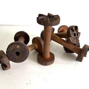 May include: A collection of six antique wooden spools. Each spool has a dark brown, aged appearance with a cylindrical body and a decorative, carved top. The spools are arranged on a white surface.