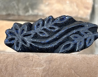 May include: A black wooden block printing stamp with a carved design of a flower and leaves. The design is outlined in blue.
