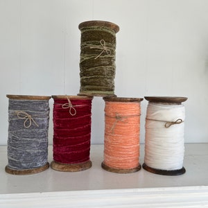 May include: Five spools of velvet ribbon on wooden spools. The ribbon colours are olive green, grey, red, peach, and white.