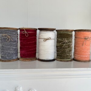 May include: Five spools of velvet ribbon in various colours, including grey, red, white, green, and orange. The spools are made of wood and have a rustic look.