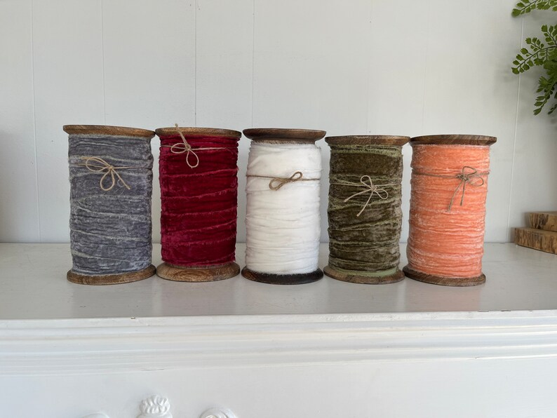 May include: Five spools of velvet ribbon in various colours, including grey, red, white, green, and orange. The spools are made of wood and have a rustic look.