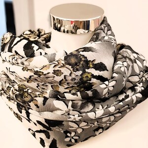 Puede incluir: Una bufanda de patrón floral blanco y gris con detalles negros y dorados. La bufanda está doblada y drapeada sobre el cuello de un maniquí.