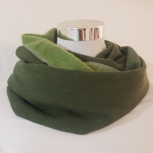 Könnte beinhalten: Ein doppellagiger Infinity-Schal mit einem dunkel olivgrünen Außenstoff und einem helleren lindgrünen Innenfutter. Der Stoff hat eine weiche, matte Textur und ist in lockeren, kreisförmigen Falten um eine weiße Schaufensterpuppe drapiert.