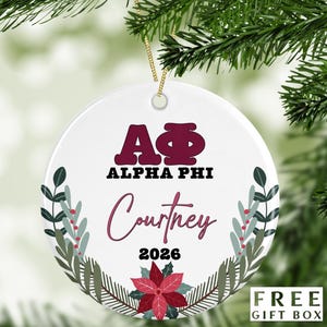 May include: A white round Christmas ornament with the text "ALPHA PHI" and "Courtney 2026" in maroon and pink. The ornament is decorated with floral and leaf designs and includes a gold string for hanging. The words "FREE GIFT BOX" are in the bottom right corner.