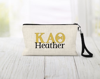 May include: A beige canvas zippered pouch with a black zipper and a black strap. The pouch has the letters "KAΘ" in gold and the word "Heather" in black.