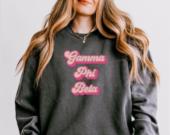 May include: Dark gray sweatshirt with the text "Gamma Phi Beta" in pink and light pink retro font. The sweatshirt has a crew neck and long sleeves. The text is stacked vertically.