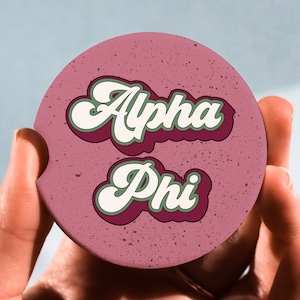 May include: Pink round coaster with the text "Alpha Phi" in white and green letters with a burgundy outline.