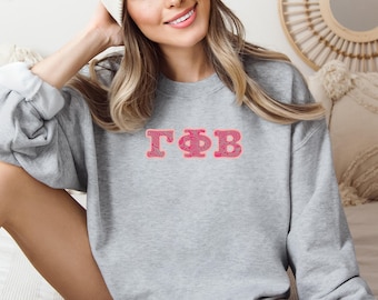 May include: A heather gray sweatshirt with the Greek letters ΓΦB embroidered in pink. The letters have a textured design. The sweatshirt has long sleeves and a crew neck. The model is wearing a white beanie.