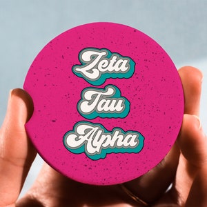 May include: A pink round coaster with the words "Zeta Tau Alpha" in white and teal lettering.