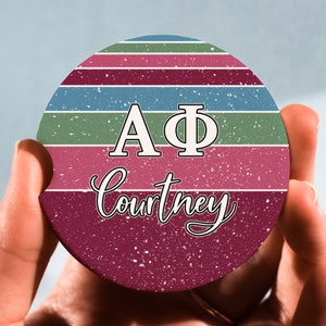 May include: A round, colorful coaster with a striped design in shades of green, blue, and burgundy. The letters "AΦ" are printed in white on the coaster, along with the name "Courtney" in a cursive font.