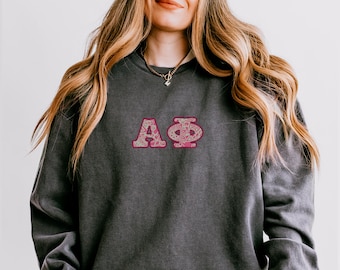 May include: Dark gray sweatshirt with the Greek letters ΑΦ in a pink floral design. The letters are embroidered on the front of the shirt. The sweatshirt has a crew neck and long sleeves.