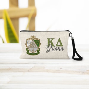 May include: A beige canvas makeup bag with a black zipper and a black strap. The bag features a green and gold embroidered crest with the letters "KA" and the name "Briana" in green.