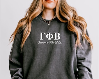 May include: Dark gray sweatshirt with white Greek letters and the words "Gamma Phi Beta" in a script font. The sweatshirt is a solid color and has a crew neck. The image shows the upper body of a person.