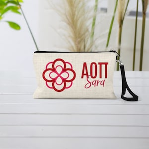 May include: A white canvas makeup bag with a black zipper and a black strap. The bag has a red and pink flower design and the text "AOT Sara" in red.