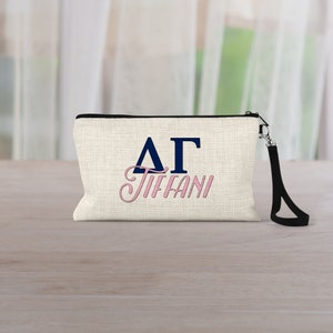 May include: A white canvas zippered pouch with a black zipper and a black strap. The pouch has blue Greek letters "ΔΓ" and the name "TIFFANI" in pink cursive lettering.