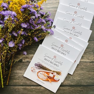 Puede incluir: Una pila de libros blancos con el título "Science of Essentials Best Recipes" en la portada. El libro presenta un diseño floral y el texto "La guía definitiva para recetas de aceites esenciales para tu mente, cuerpo y hogar".