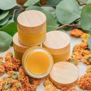 Puede incluir: Varios frascos pequeños de vidrio transparente con tapas de madera, llenos de un bálsamo amarillo. Flores naranjas secas y hojas verdes rodean los frascos, creando una estética natural y orgánica. Probablemente un producto para el cuidado de la piel.