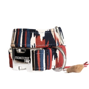 Peut inclure: Un collier de chien à motif rouge, bleu et blanc avec une boucle argentée et un pompon brun. Le collier porte le texte "FRIMOUSSE DOG" sur la boucle.