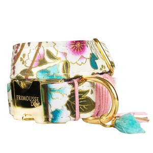 Puede incluir: Dos collares para perros apilados. Tienen un patrón floral rosa, azul y dorado sobre un fondo blanco. Los herrajes son dorados. Uno de los collares tiene una borla rosa y una borla turquesa.