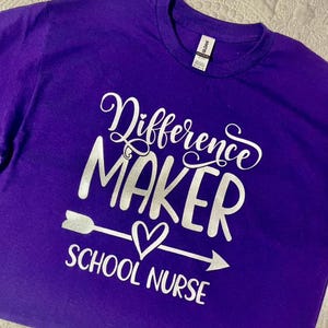 Op de afbeelding: Paars T-shirt met een zilveren glitterontwerp met de tekst "Difference Maker School Nurse" met een pijl en een hart.