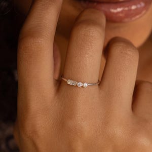 Puede incluir: Un delicado anillo de plata con tres pequeñas piedras preciosas redondas y transparentes. El anillo se lleva en un dedo, mostrando su diseño minimalista y su brillo sutil. La delgada banda del anillo y la disposición de las piedras preciosas crean una estética refinada y elegante.