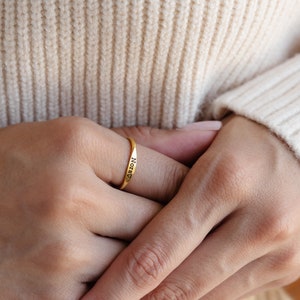 May include: A gold ring with the word "Nora" engraved on it. The ring is worn on the index finger of a person's hand.