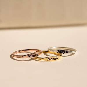 Könnte beinhalten: Drei personalisierte Ringe mit den Namen "Joey", "Nora" und "Peanut" eingraviert. Die Ringe sind übereinander gestapelt, wobei der silberne Ring oben, der goldene Ring in der Mitte und der roségoldene Ring unten liegt.