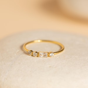 May include: A delicate gold ring featuring three round and one rectangular clear gemstones. The ring is set on a light-colored, textured surface, with a soft, neutral background. The ring's design is minimalist and elegant.