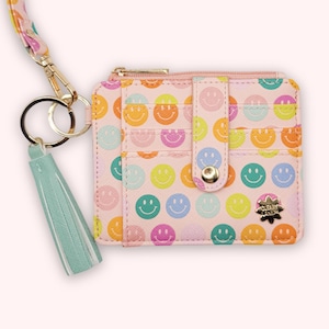 May include: Pink cardholder with a repeating pattern of colorful smiley faces. The wallet has a gold zipper, a snap closure, and a mint green tassel. A gold keychain and a patterned strap are attached.