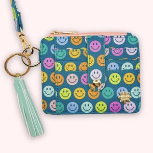 May include: Teal cardholder with a repeating smiley face pattern in various colors. The wallet has multiple card slots, a zipper closure, and a gold-tone flower charm. A matching lanyard and a mint green tassel are attached.