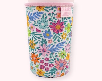 May include: A floral print drink sleeve with a pink rim and the text "CLEARY LANE". The sleeve features a colorful pattern of flowers in shades of pink, yellow, blue, and orange on a white background. The sleeve is designed to fit around a cup or bottle.