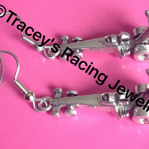 May include: A pair of silver earrings featuring a detailed miniature race car design. The earrings are on a silver hook style.