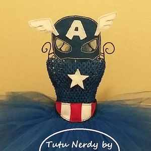 Op de afbeelding: Een Captain America-thema tutu jurk met een blauw lijfje met een witte ster en een rood-wit gestreepte kraag. De blauwe tule rok is vol en pluizig. Een Captain America-masker met witte vleugels en de letter "A" staat erop. De tekst "Tutu Nerdy by LanYapCrafts" is zichtbaar.