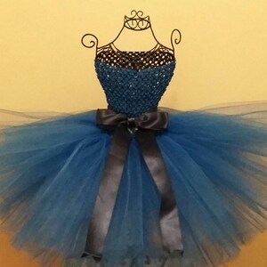 Op de afbeelding: Een blauwe tutu-jurk tentoongesteld op een draadmodel. Het lijfje is donkerblauwgroen en de rok is van gelaagd blauw tule. Een donkergrijze satijnen strik accentueert de taille. De jurk is ontworpen voor een kind.