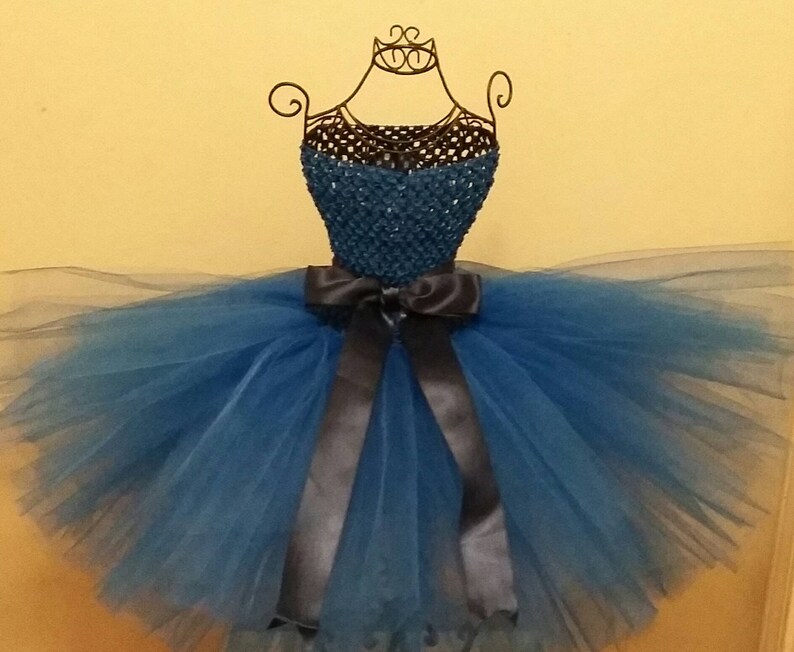 Op de afbeelding: Een blauwe tutu-jurk tentoongesteld op een draadmodel. Het lijfje is donkerblauwgroen en de rok is van gelaagd blauw tule. Een donkergrijze satijnen strik accentueert de taille. De jurk is ontworpen voor een kind.
