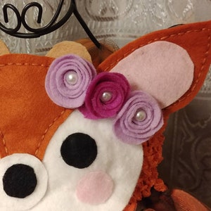 May include: A close-up the deer applique with a white face and red-orange fur. The deer has a black nose, pink cheeks, black eyes, and a crown of three felt flowers in shades of purple and pink.
