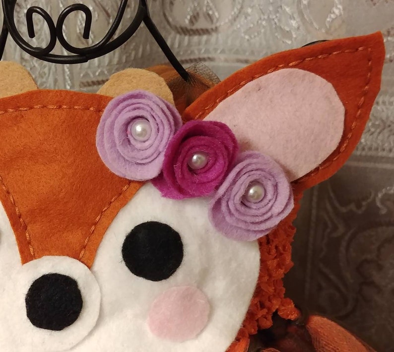 May include: A close-up the deer applique with a white face and red-orange fur. The deer has a black nose, pink cheeks, black eyes, and a crown of three felt flowers in shades of purple and pink.