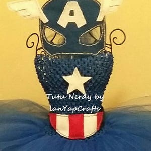 Op de afbeelding: Een Captain America-thema kostuumset, met een marineblauw masker met een witte "A" en vleugelaccenten, een blauwe lijfje met een witte ster en een rood-witte riem. Het kostuum wordt getoond op een draadstandaard, met een blauwe tutu rok.