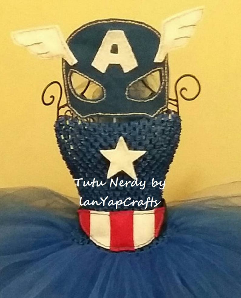 Op de afbeelding: Een Captain America-thema kostuumset, met een marineblauw masker met een witte "A" en vleugelaccenten, een blauwe lijfje met een witte ster en een rood-witte riem. Het kostuum wordt getoond op een draadstandaard, met een blauwe tutu rok.