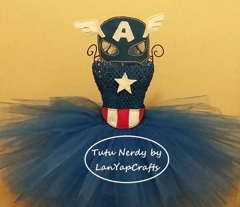 Op de afbeelding: Een Captain America-thema tutu jurk met een blauw lijfje met een witte ster en een rood-wit gestreepte kraag. De blauwe tule rok is vol en pluizig. Een Captain America-masker met witte vleugels en de letter "A" staat erop. De tekst "Tutu Nerdy by LanYapCrafts" is zichtbaar.
