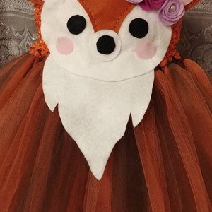 May include: A brown tulle tutu with a deer-shaped felt applique. The deer has pink ears, a white face, black eyes, and a black nose. The deer is decorated with three purple felt flowers with crystal gem centers.