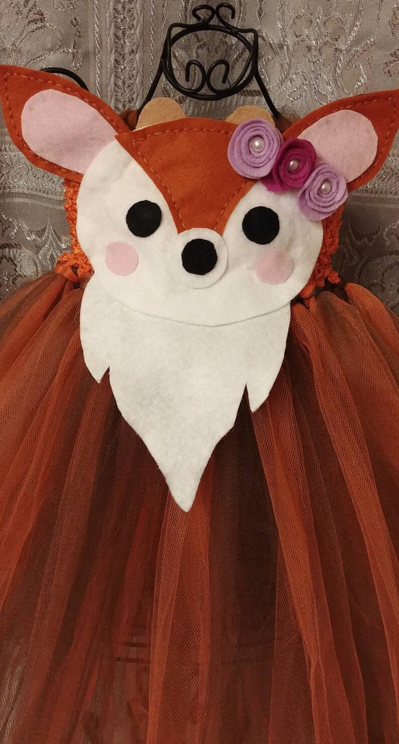 May include: A brown tulle tutu with a deer-shaped felt applique. The deer has pink ears, a white face, black eyes, and a black nose. The deer is decorated with three purple felt flowers with crystal gem centers.