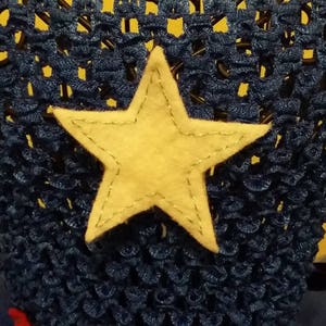 Op de afbeelding: Een donkerblauwe gehaakte hoofdband met een grote, lichtgele sterapplicatie. De ster is op de hoofdband genaaid. De hoofdband is waarschijnlijk voor een kind en staat tegen een gele achtergrond.