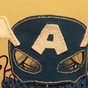 Op de afbeelding: Een blauw vilten masker met witte vleugels en een witte "A" in het midden. Het masker heeft oogopeningen en is afgewerkt met witte stiksels. De vleugels zijn wit met grijze stiksels. Het masker wordt op een decoratieve standaard getoond.